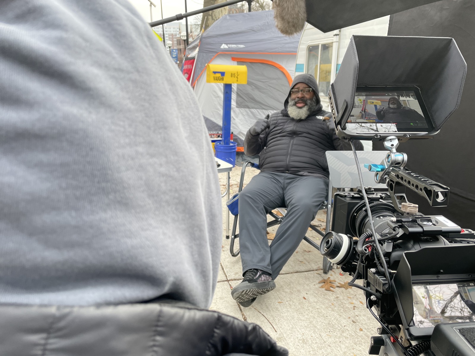 Cinema camera rig with monitor trained on Jon Vaughn in a hooded winter coat at his protest encampment during Hail To The Victims
