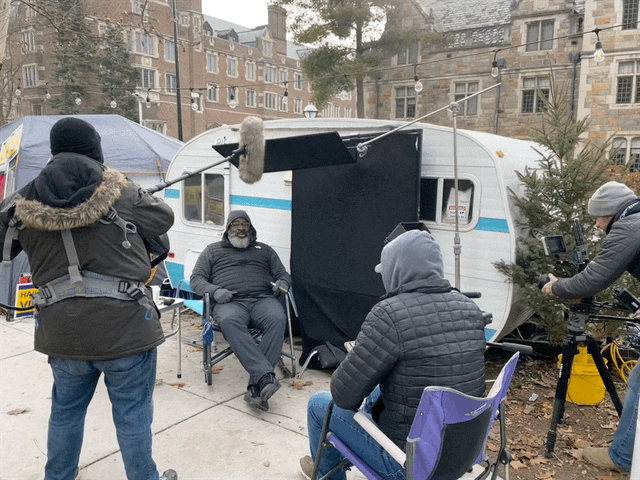 Film crew with a boom mic interviewing Jon Vaughn in front of a vintage camper on the University of Michigan campus during Hail To The Victims